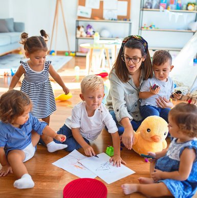 Lehrerin und eine Gruppe Kleinkinder sitzen auf dem Boden und zeichnen mit Papier und Buntstiften