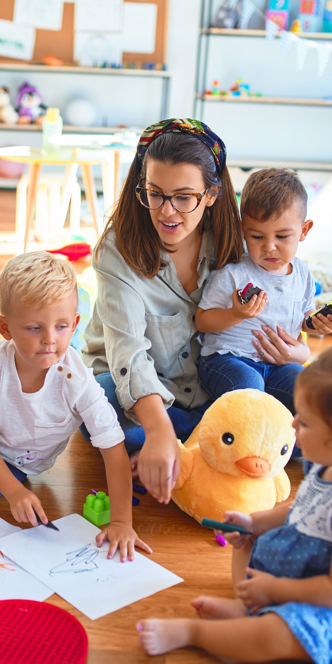 Lehrerin und eine Gruppe Kleinkinder sitzen auf dem Boden und zeichnen mit Papier und Buntstiften