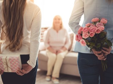 Zwei Personen halten Blumen und eine Geschenkbox hinter ihrem Rücken als Überraschung für ihre auf dem Sofa sitzende Oma