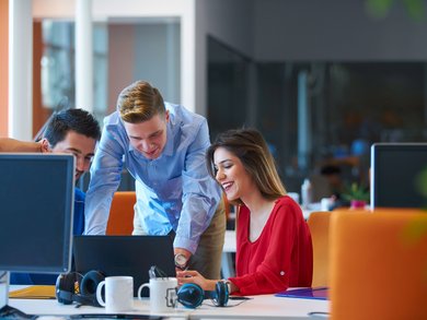 Drei Personen, die in einem Büro über eine Laptop gebeugt sind (etwas betrachten)