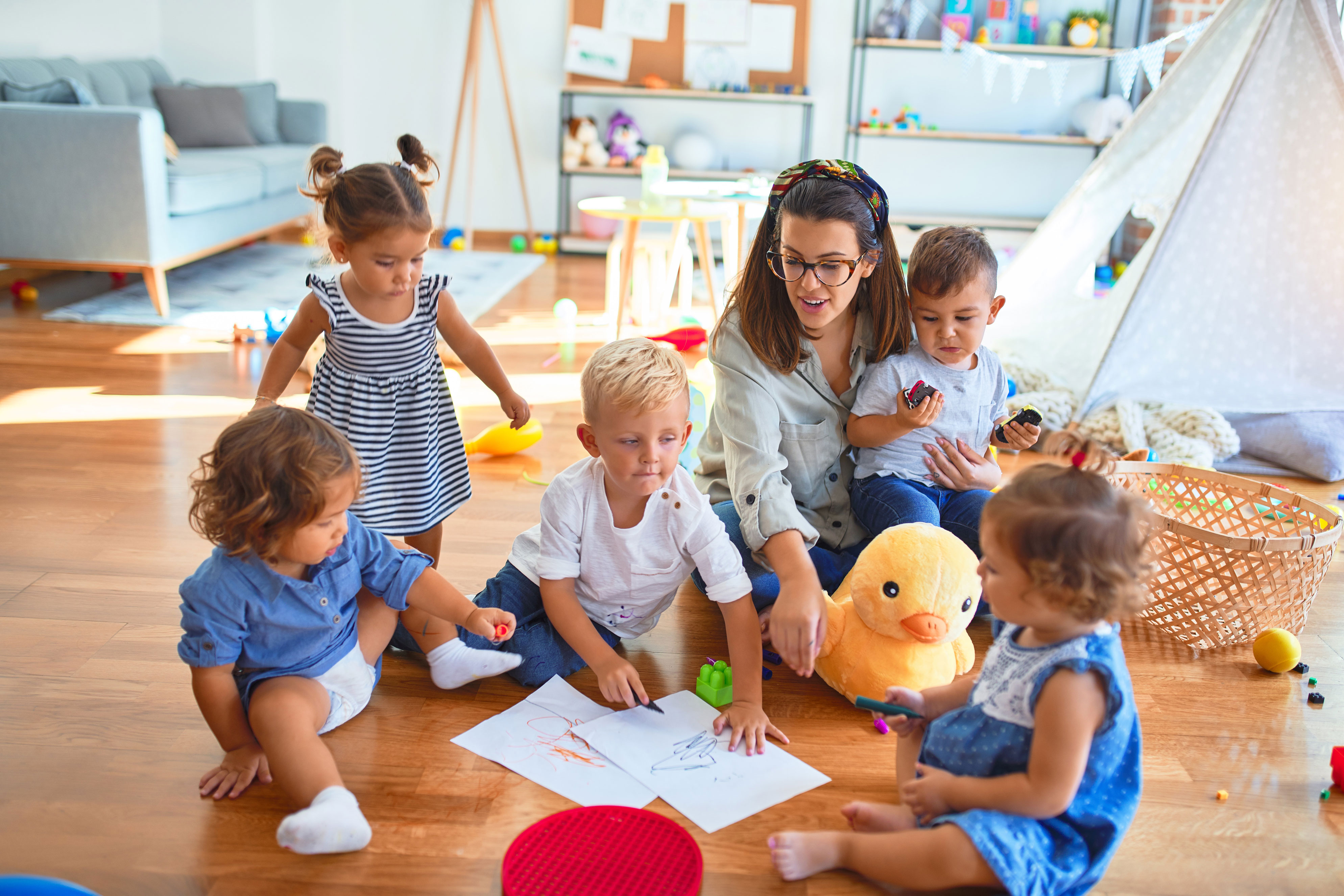 Lehrerin und eine Gruppe Kleinkinder sitzen auf dem Boden und zeichnen mit Papier und Buntstiften